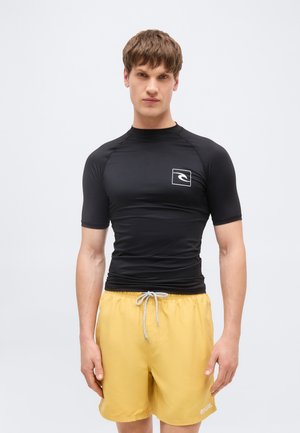 Man met een strakke zwarte kortarmige top met logo en gele zwemshort met trekkoord, staand tegen een effen lichte achtergrond.