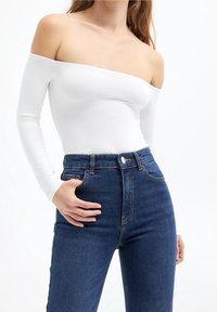 Body blanc à manches longues et épaules dénudées, associé à un jean taille haute bleu foncé avec un design classique à cinq poches et un bouton en métal.