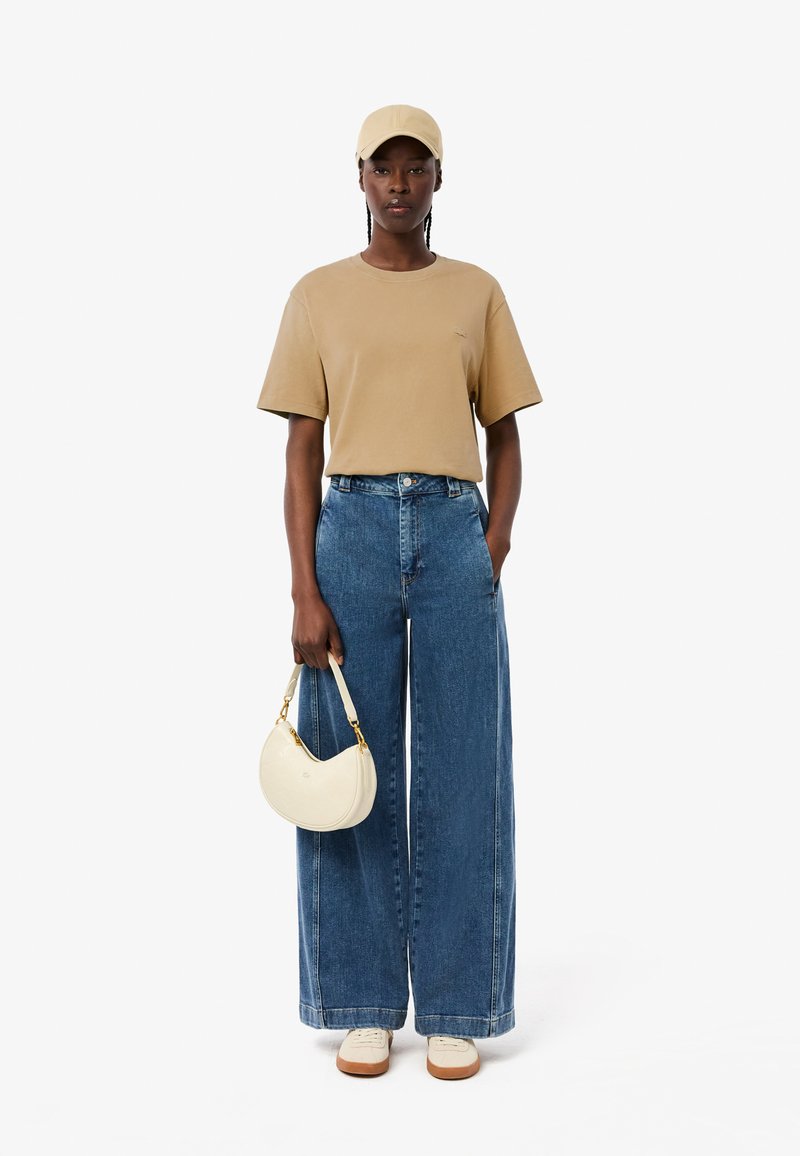 Beige T-Shirt mit kurzen Ärmeln, hochgeschnittene weite Denim-Jeans, cremefarbene runde Handtasche und beiger Cap, dazu weiße Sneakers mit braunen Sohlen.