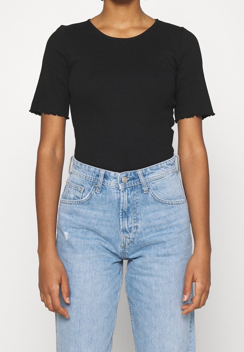 Personne portant un haut noir côtelé à manches courtes ajusté et un jean taille haute bleu clair avec poches avant et fermeture par boutons.