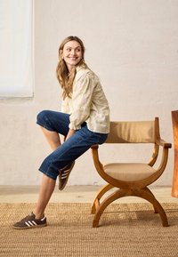 Lächelnde Frau mit langen Haaren sitzt seitlich auf einem Holzstuhl mit beigem Kissen, trägt eine helle Bluse, gekürzte Jeans und braune Turnschuhe.