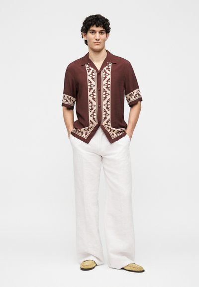 Άνδρας στέκεται με τα χέρια στις τσέπες, φοράει καφέ κοντομάνικο πουκάμισο με μπεζ σχέδια, λευκό παντελόνι με φαρδιά μπατζάκια και μπεζ παπούτσια.