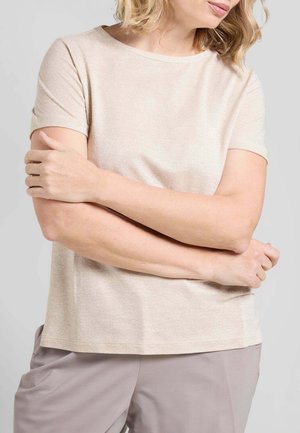 Donna con capelli biondi che indossa una maglietta beige a maniche corte e pantaloni grigio chiaro, con le braccia incrociate all'altezza della vita su uno sfondo neutro.