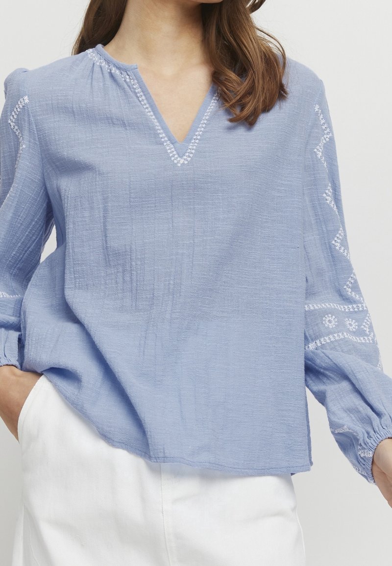 Vaaleansininen, teksturoitu pusero, jossa V-aukkoinen pääntie, kerätty yksityiskohta ja valkoiset kirjailut hihoissa. Löysä istuvuus ja joustavat hihansuut.