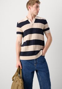 Cămașă polo tricotată în albastru închis și crem, cu un model cu dungi; are decolteu în V și mâneci scurte. Modelul ține o geantă bej.