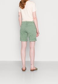 Pantalones cortos verdes con un patrón sutil, que cuentan con dobladillos enrollados y un ajuste clásico. Se llevan con una camiseta beige clara y sandalias planas.