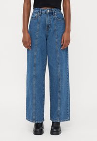 Jeans in denim blu a gamba larga con vita alta, caratterizzati da evidenti dettagli di cucitura e tasche frontali. Indossati con stivali neri alla caviglia.