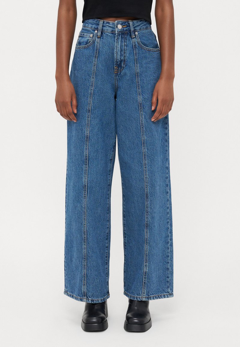 Jeans in denim blu a gamba larga con vita alta, caratterizzati da evidenti dettagli di cucitura e tasche frontali. Indossati con stivali neri alla caviglia.