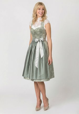 Blonde Frau trägt ein hellgrünes traditionelles Kleid mit Spitzenmieder, einer Satinschürze, die zu einer Schleife gebunden ist, und beigen High Heels, lächelnd und stehend.