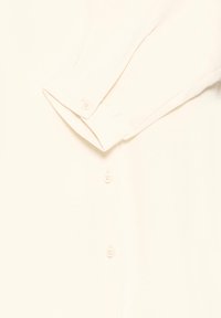 Krémová košeľa s button-up zapínaním, dlhými rukávmi a zapínaním na gombíky, a hladkou textúrou látky.