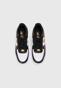 Sapatilhas de couro em preto e branco com detalhes em dourado, apresentando a biqueira perfurada, atacadores pretos e uma etiqueta dourada na língua.