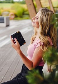 Black leather wallet with a textured surface, gold hardware accents, held by a person in a pink top, sitting in a natural outdoor setting.