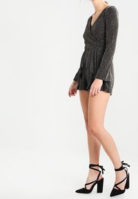 Robe enveloppante noire brillante à manches longues et à ourlet évasé, associée à des chaussures à talons hauts noires avec des brides de cheville.