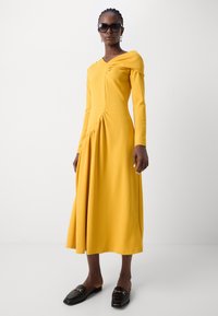 Pitkä keltainen mekko, jossa on V-aukko, rypytetty olkapäätyyli ja sivujen drapointi. Päällä mustat loafer-kengät, joissa on kultaiset yksityiskohdat.