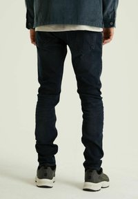 Donkerblauwe jeans met een slim fit en een subtiele vervaging. Gecombineerd met grijze en zwarte sneakers. Achtergrond is effen licht van kleur.