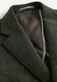 Grüne wollige Jacke mit strukturiertem Oberflächenfinish. Sichtbares graues Futter und subtile geometrische Muster im Inneren. Klassisches Reversdesign.