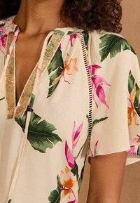 Blumenbluse mit einem cremefarbenen Hintergrund, lebhaften pinken und orangen Blumen, grünen Blättern, einem V-Ausschnitt und goldenen Paillettenakzenten.