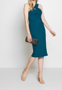 Robe midi ajustée en bleu sarcelle avec un design à une épaule, présentant une texture lisse. Associée à une pochette noire ornée de sequins et des talons beiges à lanières.