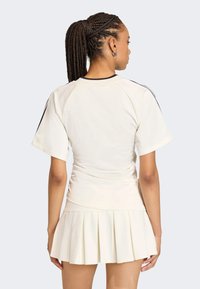 Krém színű sportos felső rövid ujjakkal és fekete részletekkel, élénkített szoknyával párosítva. Az anyag sima, testhez simuló szabású.