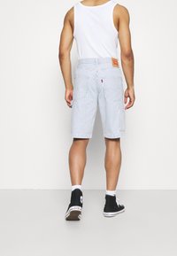 Man i vit linne, blå och vita randiga shorts, vita strumpor och svarta hög-top sneakers som står vänd bort mot en neutral bakgrund.