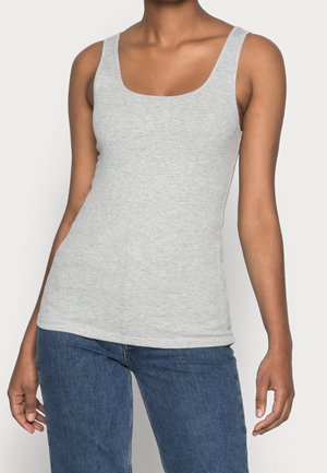 Person trägt ein enganliegendes hellgraues, geripptes Tanktop und blaue Jeans, steht vor einem schlichten hellen Hintergrund.