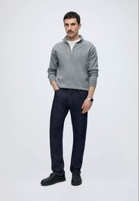Mand med overskæg iført grå halvlange lynlås sweater, mørke jeans, sorte sko og ur, stående mod en ensfarvet lys baggrund.