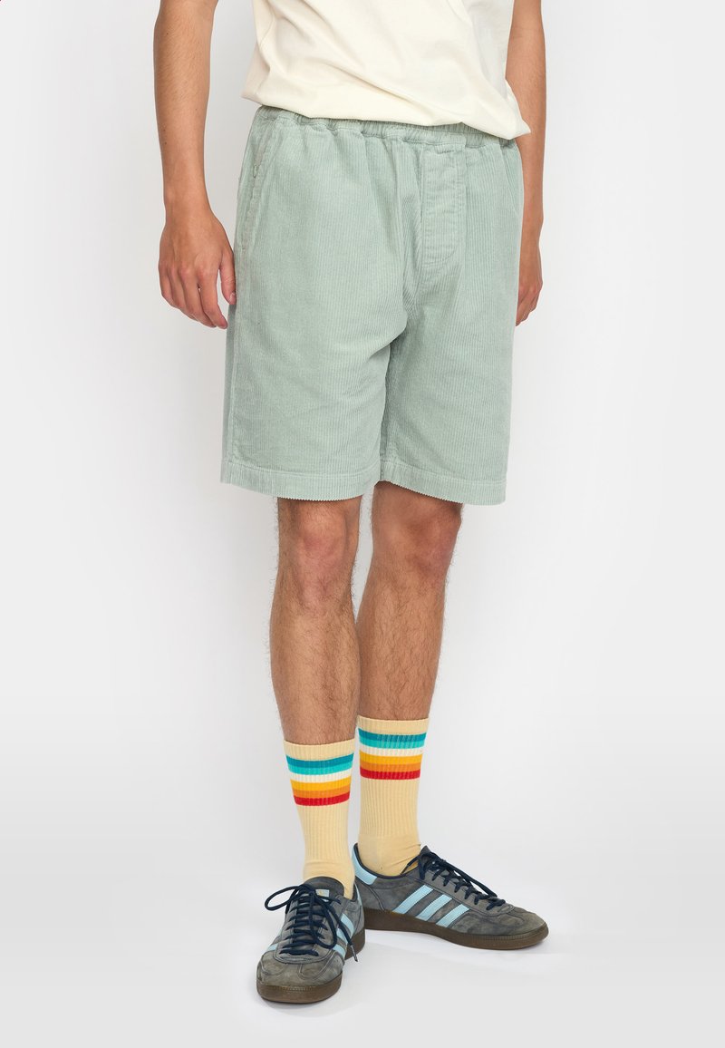 Lichtgroene corduroy short met een elastische tailleband, met zijzakken. Draagt bij multicolore gestreepte sokken en donkere sneakers.