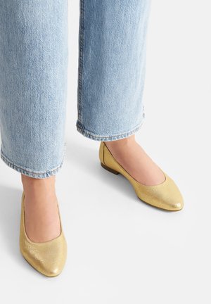 Złote, teksturowane baleriny z zaokrąglonym noskiem i niskim obcasem, zestawione z jasnoniebieskim denimem z surowym wykończeniem na dole.