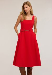 Rochie roșie fără mâneci, cu decolteu pătrat, accesorizată cu un brâu din lanț la talie, având buzunare laterale și o fustă evazată.