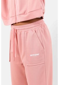 Conjunto de chándal rosa que incluye una sudadera recortada y pantalones a juego. Fabricado con un tejido suave con un acabado texturizado. Logo en la pierna del pantalón.