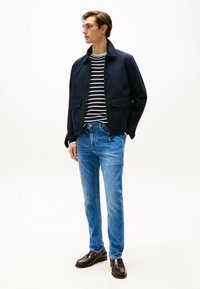 Giacca bomber navy sopra una maglietta a righe bianche e nere, abbinata a jeans azzurri chiari e mocassini marrone scuro. Tessuto morbido e vestibilità casual.