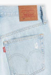 Light blue denim jeans featuring a brown leather patch with embossed logo at the waistline and a red tag on the back pocket.
