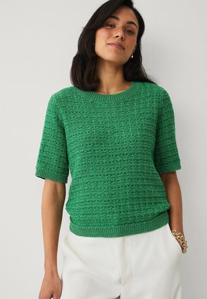 Vrouw draagt een gestructureerde groene trui met korte mouwen en witte broek, staand met één hand in de zak tegen een effen achtergrond.