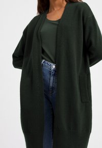 Mörkgrön stickad cardigan med långa ärmar, öppen fram, och en ficka på vänster sida, kombinerad med en mörkgrön topp och blå denimjeans.