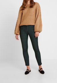 Suéter de punto grueso en tono tostado con un patrón de trenzas, combinado con jeans ajustados verde oscuro y zapatos planos negros con una correa.