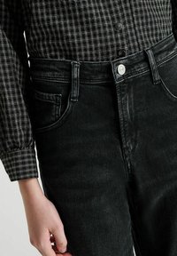 Close-up van een persoon die zwart hoge taille jeans draagt met een knop en riemlussen, gecombineerd met een zwart-wit geruit shirt.