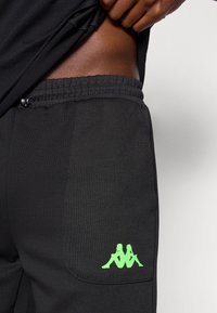 Fekete jogger nadrág elasztikus derékpánttal és húzózsinórral, valamint oldalzsebbel, a hátulján zöld Kappa logóval díszítve.