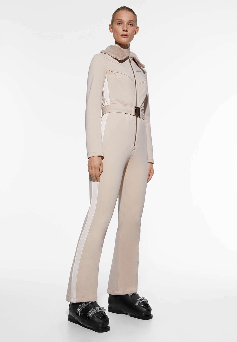 Mono cremoso con cremallera, paneles laterales blancos en contraste y un cuello de piel sintética. Presenta una cintura con cinturón y pantalones acampanados. Botas negras completan el look.