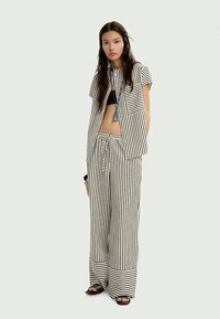 Donna che indossa una camicia a maniche corte a righe verticali bianche e nere e pantaloni a gamba larga, sandali neri e una collana con pendente lungo, in piedi.
