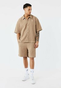 Camisa corta de manga corta con textura marrón y botones en la parte delantera, combinada con pantalones cortos a juego. Zapatillas blancas y calcetines con estampado completan el conjunto.