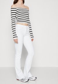 Un crop top noir et blanc à rayures, dégageant les épaules, associé à un jean évasé blanc et des chaussures de sport argentées. Textures de tissu lisses.