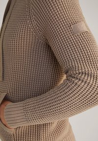 Beige gestrickter Pulloverärmel mit Waffelmuster, geripptem Bündchen und einem Stoffmarkenpatch am Oberarm; Hand teilweise in der Tasche.