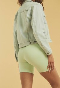 Vaaleansininen farkkutakki, jossa on kulunut pinta, yhdistettynä mintunvihreisiin pyöräilyshortseihin. Keskiössä takin hienovarainen tekstuuri ja nappidetalit.