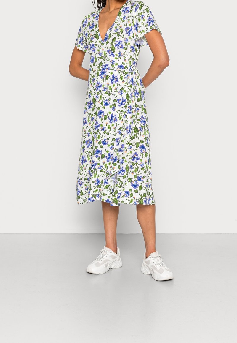 Femme portant une robe midi portefeuille blanche avec un imprimé floral bleu et des manches courtes, associée à des baskets blanches, debout contre un mur uni.