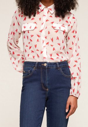 Femme portant un chemisier blanc transparent avec des motifs de cœurs rouges, rentré dans un jean bleu foncé taille haute, debout avec une main détendue.