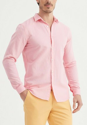 Homme portant une chemise rose clair à boutons et un pantalon jaune, debout avec une main dans la poche devant un fond uni.