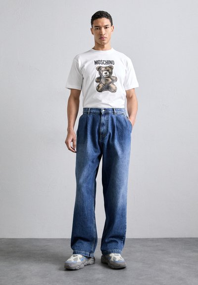 T-shirt en coton blanc avec un graphique d'ours brun et le texte "MOSCHINO", associé à un jean en denim bleu ample et des baskets grises.