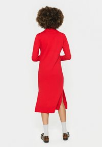 Abito rosso a maglia con colletto alto, maniche lunghe e spacco laterale. Presenta una texture liscia e una silhouette aderente.