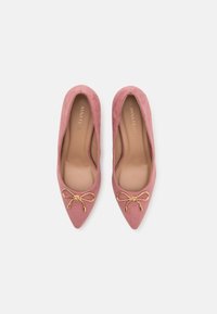Paire de ballerines pointues en daim rose avec des accents de nœud doré et des semelles intérieures beiges, vues de dessus sur un fond blanc.