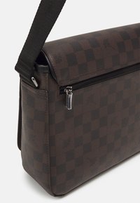 Bolso mensajero marrón a cuadros con un acabado texturizado, que cuenta con un bolsillo con cremallera y una correa ajustable. Diseño compacto y rectangular.